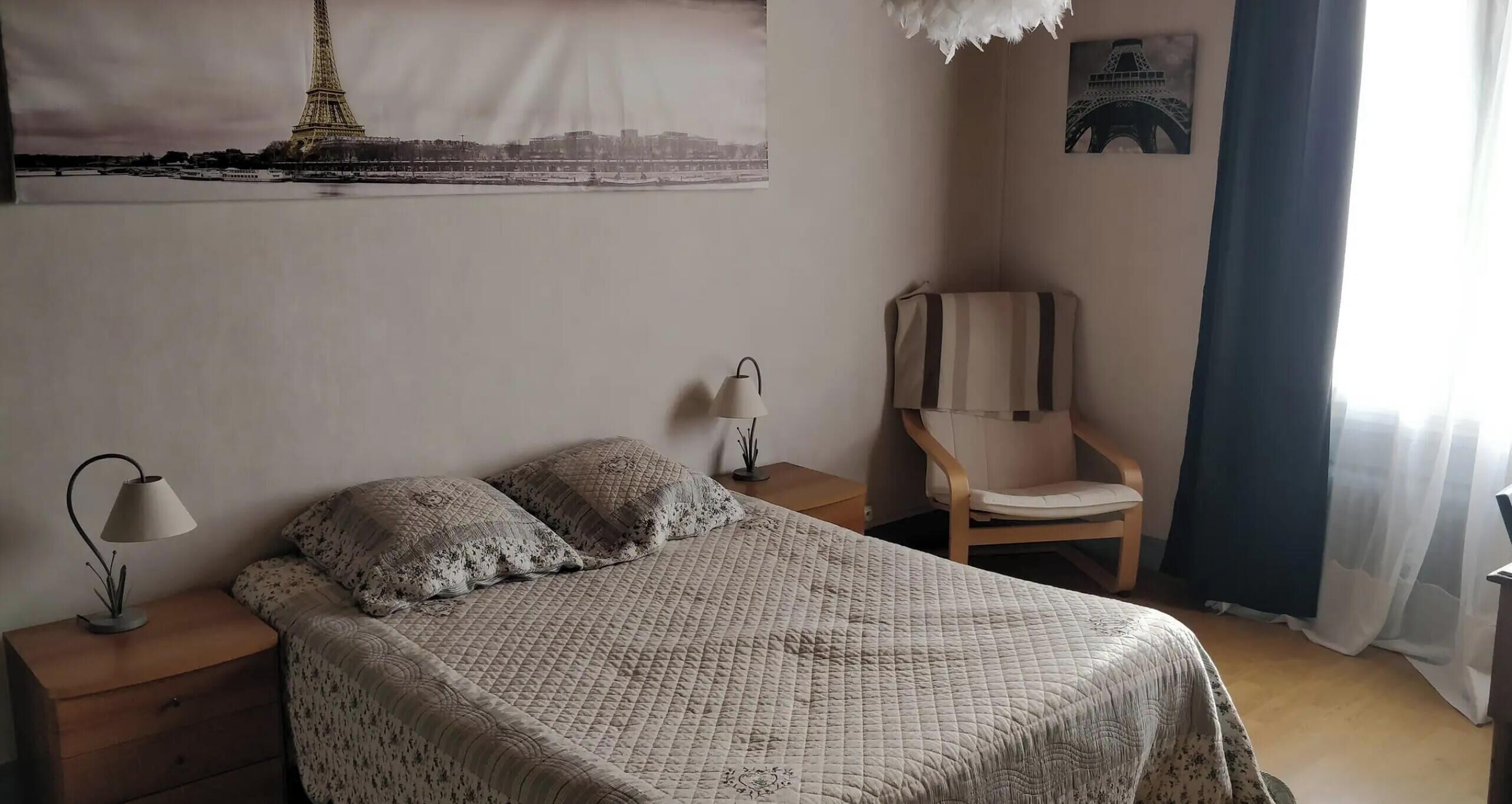A bedroom with a bed, a chair, a nightstand, a lamp, and a picture of the Eiffel Tower on the wall.