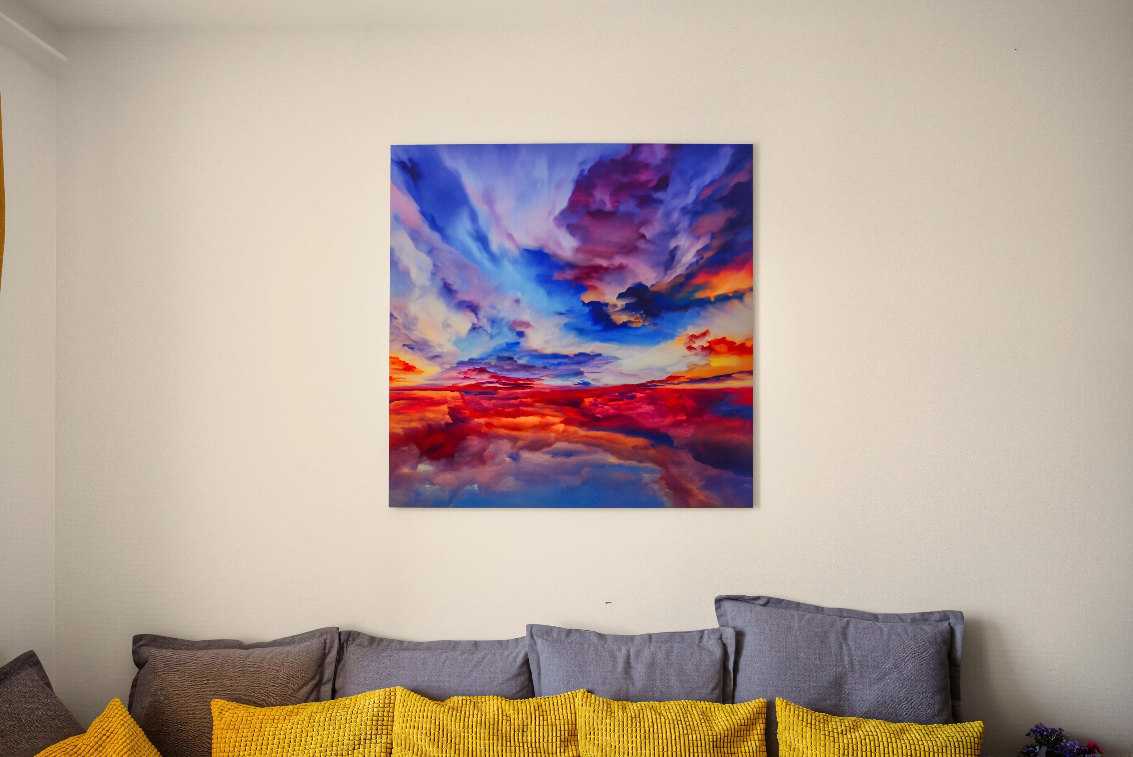 Een kamer met één kunstwerk aan de muur, een bank met kussens, en een kleine plant aan de zijkant.
