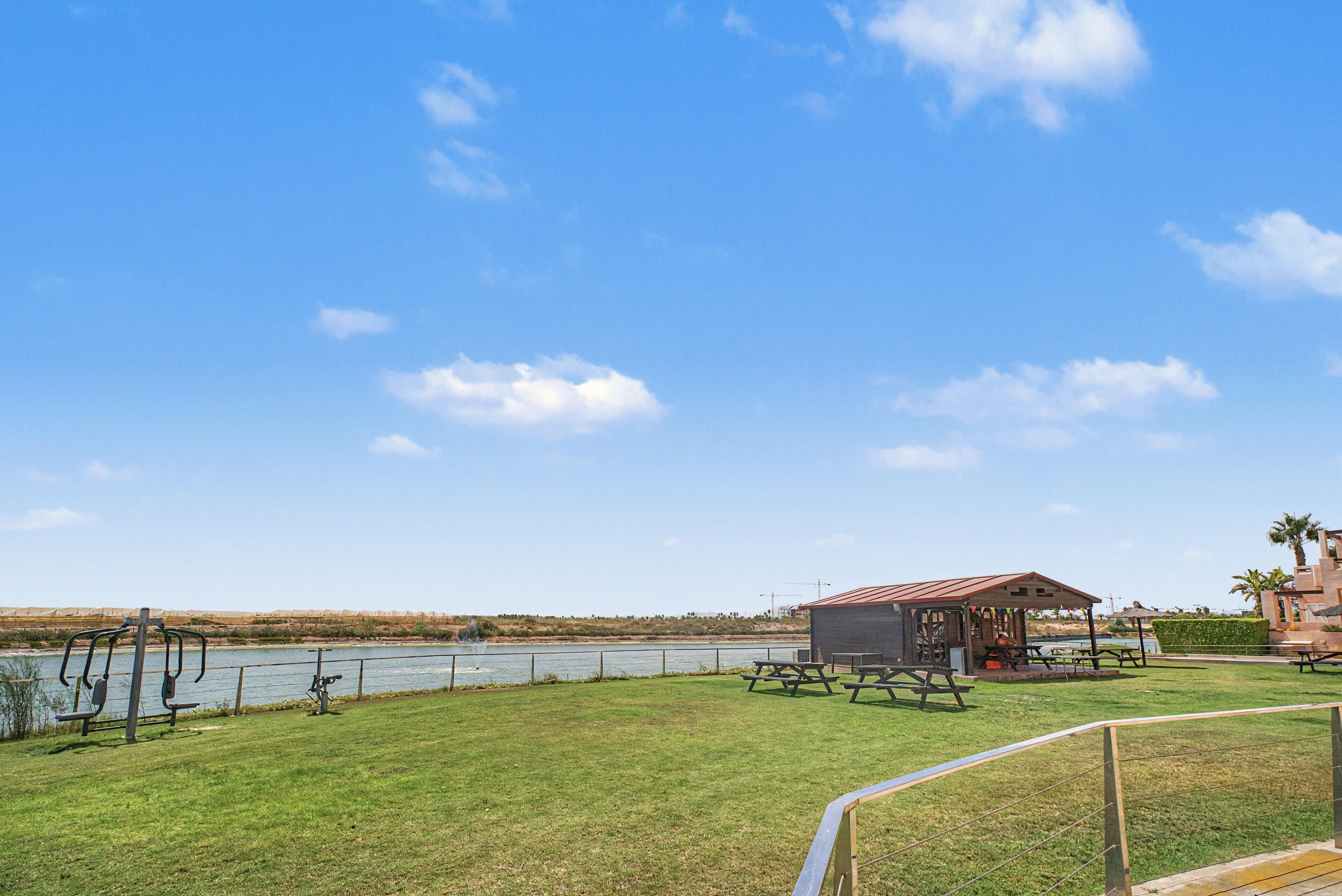 A grassy area with a small pavilion, picnic tables, and exercise equipment by a body of water.