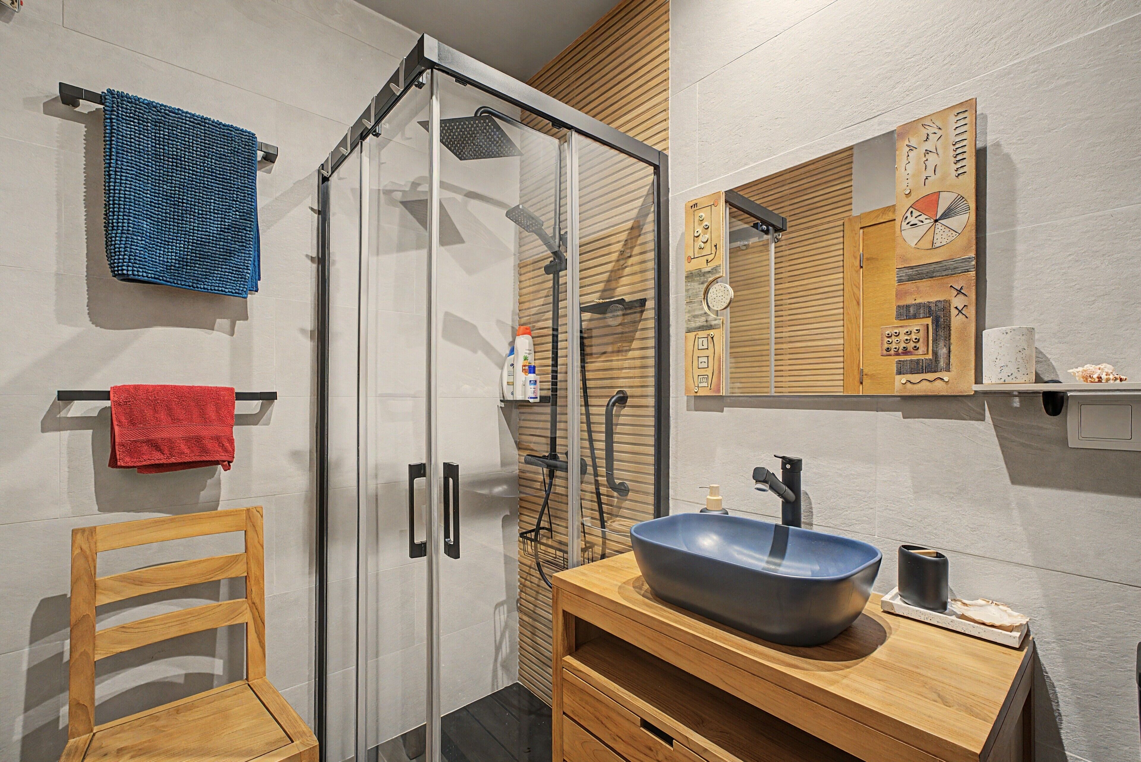 A modern bathroom with a glass shower enclosure, a wooden vanity with a blue sink, and a mirror with a decorative wooden panel.