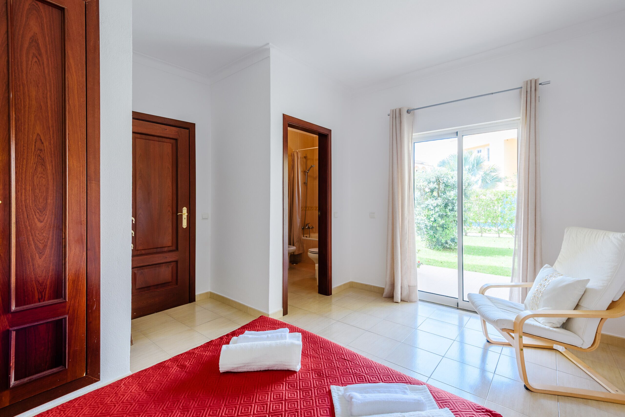 A hotel room with a red patterned carpet, a white chair, a wooden door, and a view through a glass door.