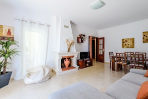 A living room with a fireplace, a sofa, a dining table with chairs, and a bookshelf. - Vilamoura Garden and Pool Apartment (Loulé)