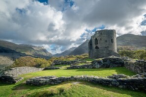 Eine historische Steinarchitektur mit Ruinen und einem grünen Feld, umgeben von Bergen und einem bewölkten Himmel.