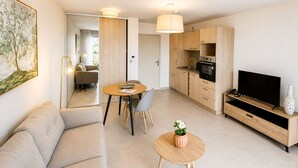 A modern living room with a sofa, a dining table with chairs, a kitchen with wooden cabinets, and a TV on a wooden stand.