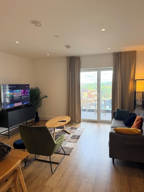 A modern living room with a flat-screen TV, a sofa, a green chair, a wooden coffee table, and a view of the outdoors through large windows.
