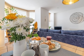 Ein Esstisch mit einem Blumenstrauß, einer Kaffeekanne, einer Tasse, einem Teller mit Croissants und einer Schüssel Orangen.