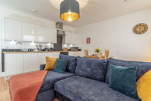 Et moderne køkken med hvide skabe, et spiseområde med et bord og stole, samt en stue med en sofa og puder.