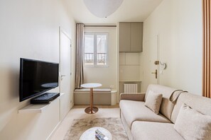 A compact living space with a flat-screen TV, a small round table, a sofa, and a window with curtains. - Montmartre-Mobility lease-Studio-2 persons-Renovated (Paris)