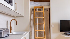 Eine kompakte Küche mit Mikrowelle, Toaster und Spüle. Eine Holzleiter lehnt an einem Vorhang, und ein Fernseher ist an der Wand montiert.