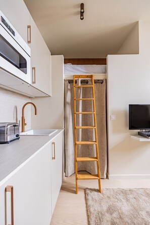 A compact kitchen with a microwave, toaster, and sink. A wooden ladder is placed against a curtain, and a TV is mounted on the wall. - Montmartre-Mobility lease-Studio-2 persons-Renovated (Paris)