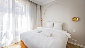 Un letto rifatto con lenzuola bianche e due asciugamani, una grande finestra con tende leggere e un lampadario a parete.