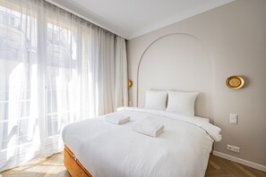 A neatly made bed with white linens and two towels, a large window with sheer curtains, and a wall-mounted light fixture. - Premium-New mobility lease-Paris16th-2bedroom (Paris)