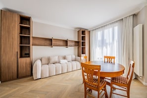 A modern living room with a sofa, wooden dining table, chairs, built-in wooden shelving, and a radiator. - Premium-New mobility lease-Paris16th-2bedroom (Paris)