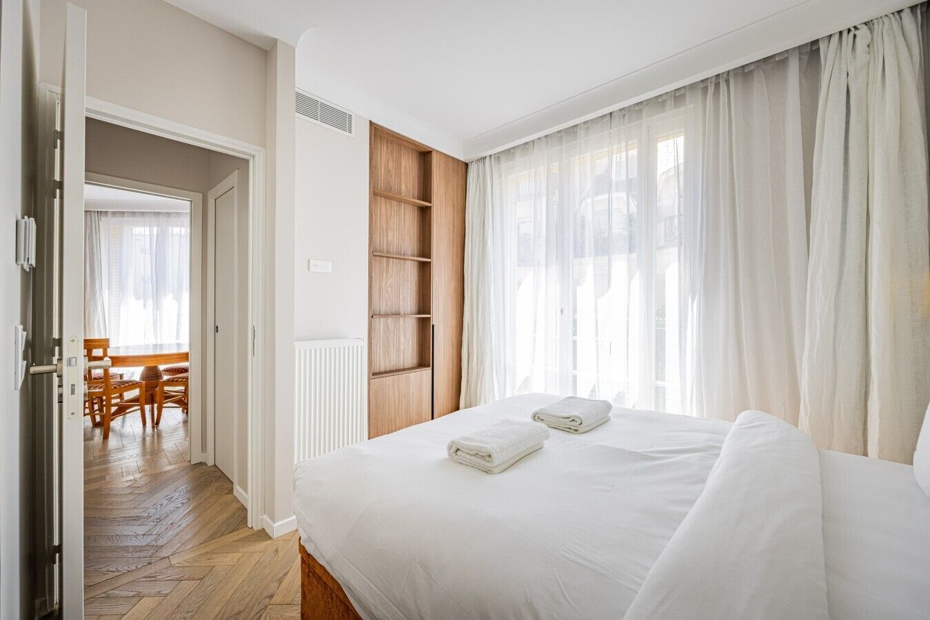 A bedroom with a bed, a wooden wardrobe, a window with sheer curtains, and a view of a dining area.