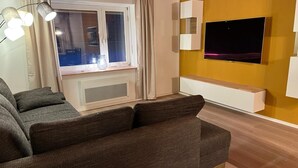 A living room with a sofa, a TV mounted on the wall, a window with curtains, and a radiator. - Our small chalet in the heart of the Palatinate Forest. Relaxation, hiking, romance. (Lindenberg)
