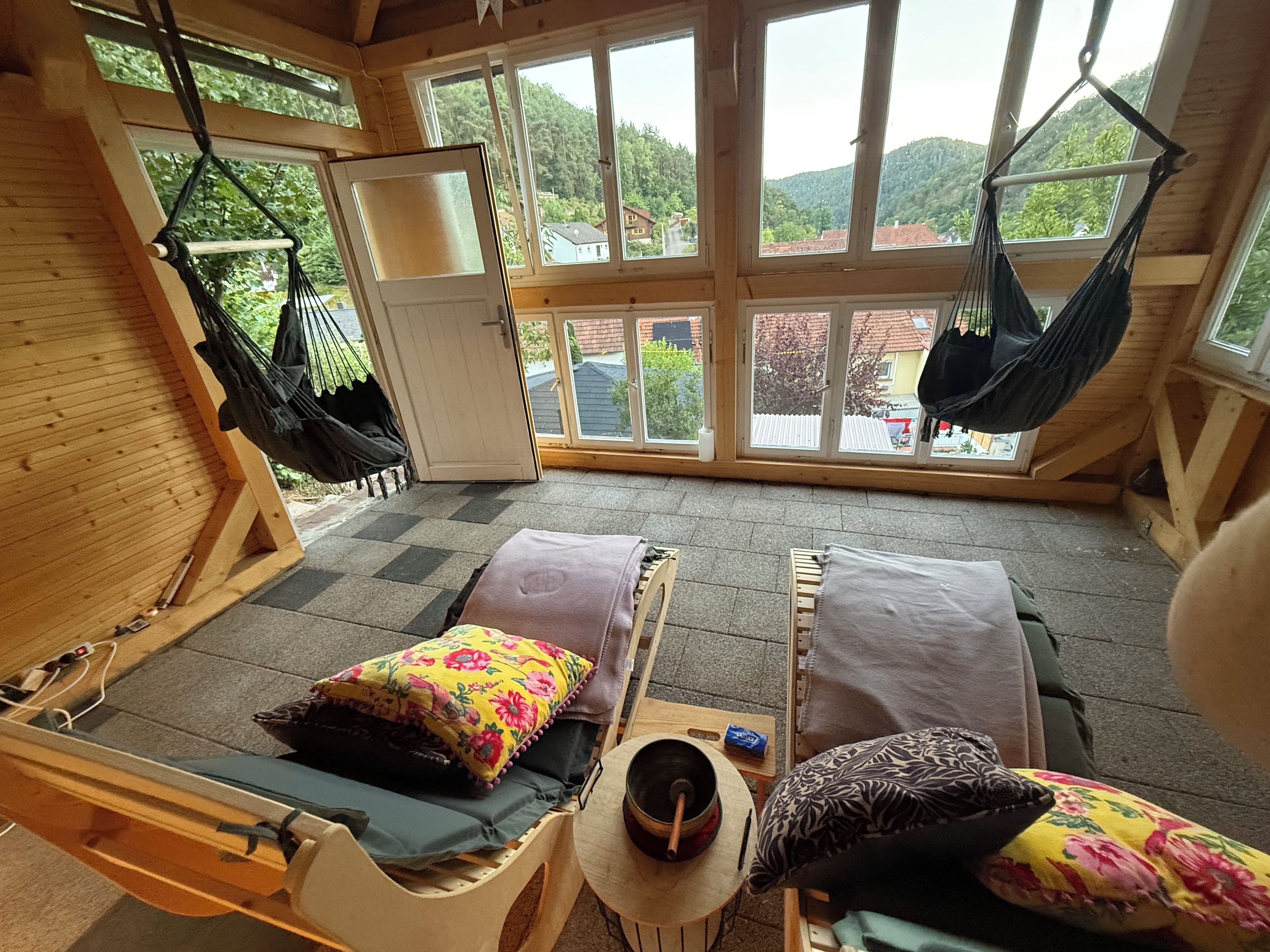 A room with two hammocks, a bed with floral pillows, and a view of a mountainous landscape through large windows.