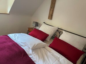 Una cama con ropa de cama blanca y roja, un cabecero de madera, una mesita de noche con una lámpara y una estantería en la pared.