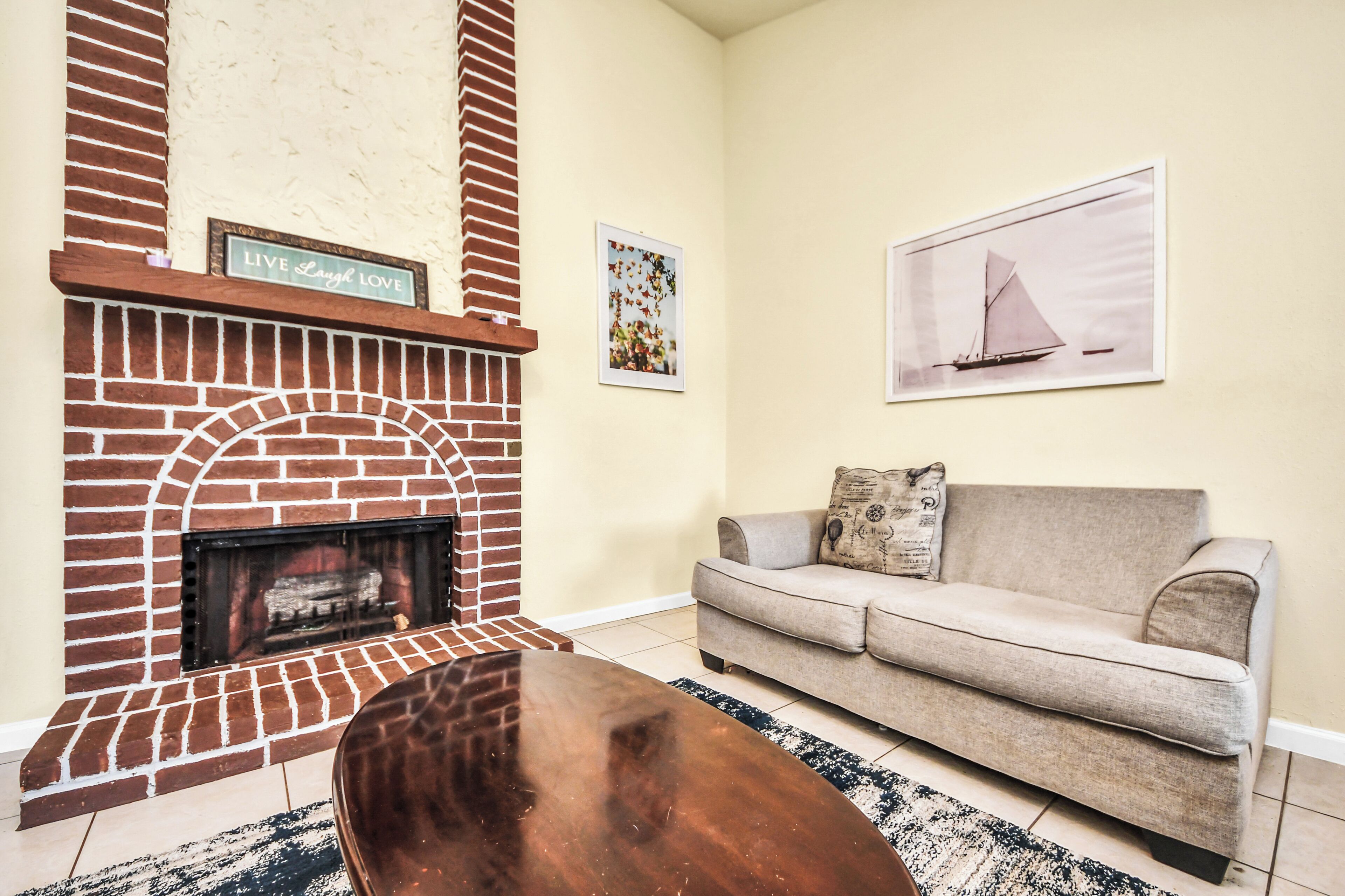 A living room with a brick fireplace, a beige sofa, a coffee table, and two framed pictures on the wall.