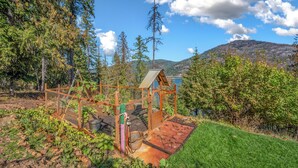 A rustic wooden structure with a metal roof, surrounded by a garden and a view of a lake and mountains. - SANDBAR LAKE CABIN- Cozy 1-bedroom cabin on 3 very private acres.  (Sandpoint)