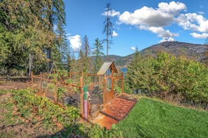 A rustic wooden structure with a metal roof, surrounded by a garden and a view of a lake and mountains.