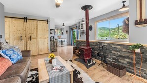 A spacious living room with a large wooden wardrobe, a red wood-burning stove, a cowhide rug, and a view of the outdoors. - SANDBAR LAKE CABIN- Cozy 1-bedroom cabin on 3 very private acres.  (Sandpoint)