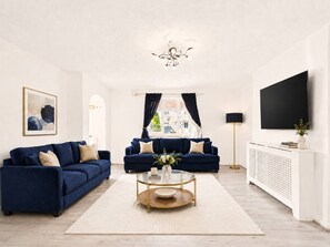 A living room with two dark blue sofas, a glass coffee table, a white radiator, and a flat-screen TV mounted on the wall. - Hatfield Retreat | Parking | Spacious | Sleeps 5 (Hertfordshire)