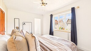 Une chambre à coucher avec un lit, des oreillers, un ventilateur de plafond, une fenêtre avec des rideaux et une vue sur les bâtiments à l’extérieur.