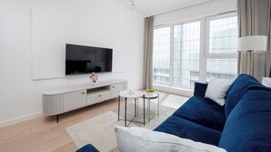Een moderne woonkamer met een flatscreen televisie, een bank, een klein rond tafeltje en een groot raam met gordijnen.