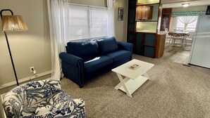 Ein Wohnzimmer mit einem blauen Sofa, einem weißen Couchtisch, einem gemusterten Sessel, einer Lampe und einer Küchenzeile inklusive Kühlschrank und Esstisch.