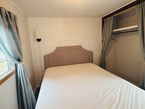 Una cama individual con cabecera, una ventana con cortinas, una lámpara de pared y un armario con espejo.