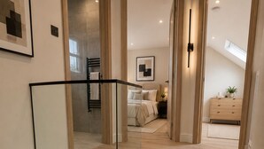 Un escalier moderne avec des rampes en verre, des marches en bois et une vue sur une chambre à coucher où l’on aperçoit un lit, une table de chevet et une commode.