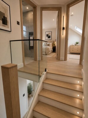 Una escalera moderna con barandillas de vidrio, escalones de madera y vistas a un dormitorio con cama, mesita de noche y cómoda.