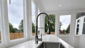 Une cuisine moderne dotée d’un évier en acier inoxydable, de plans de travail en marbre blanc et de grandes fenêtres offrant une vue sur l’extérieur.