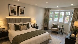 Une chambre à coucher moderne avec un grand lit, des tables de chevet, une chaise, une petite table avec une bougie, et une vue sur l’extérieur grâce à de grandes fenêtres.