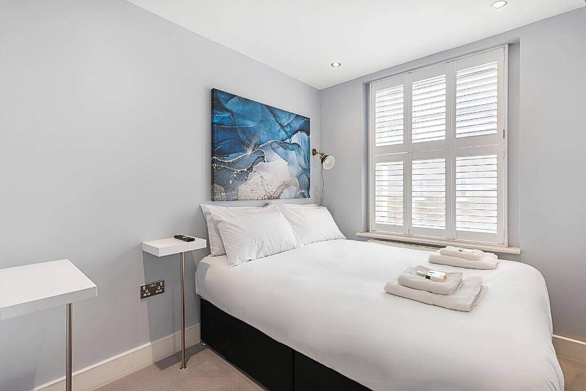 Un dormitorio moderno con cama, ropa de cama blanca, un cuadro, una mesita de noche y una ventana con persianas.