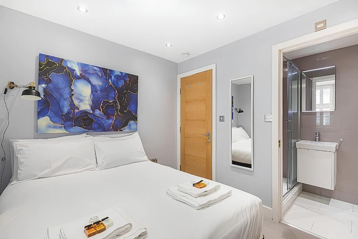 Una habitación de hotel con cama, un cuadro, una puerta de madera, un baño con espejo y lavabo.