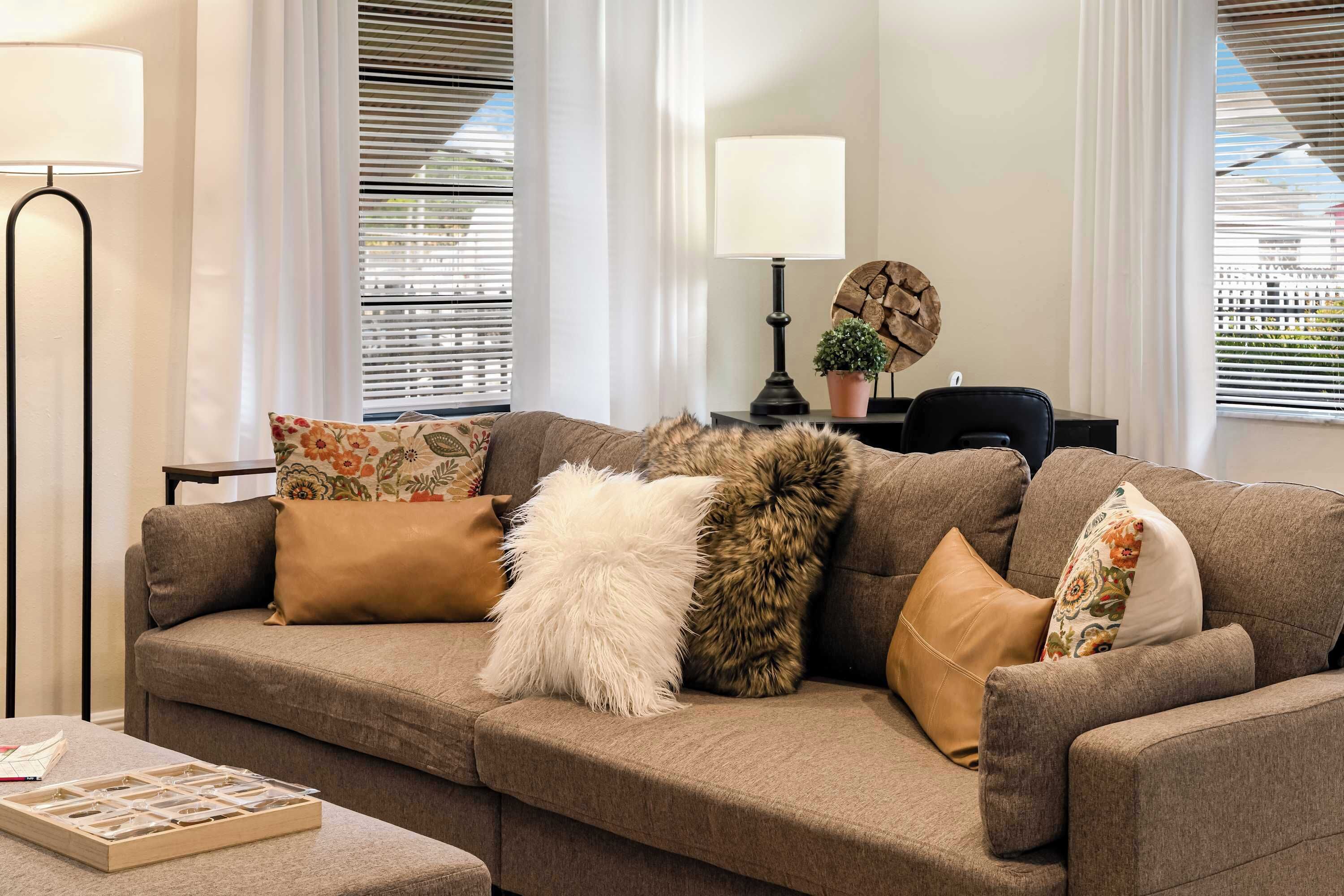 A living room with a sofa, pillows, a lamp, a small table, and a window with blinds.
