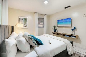 Ein ordentlich eingerichtetes Schlafzimmer mit einem Bett, Kissen, einer Nachttischlampe, einem an der Wand montierten Fernseher und einem Regal mit Büchern.