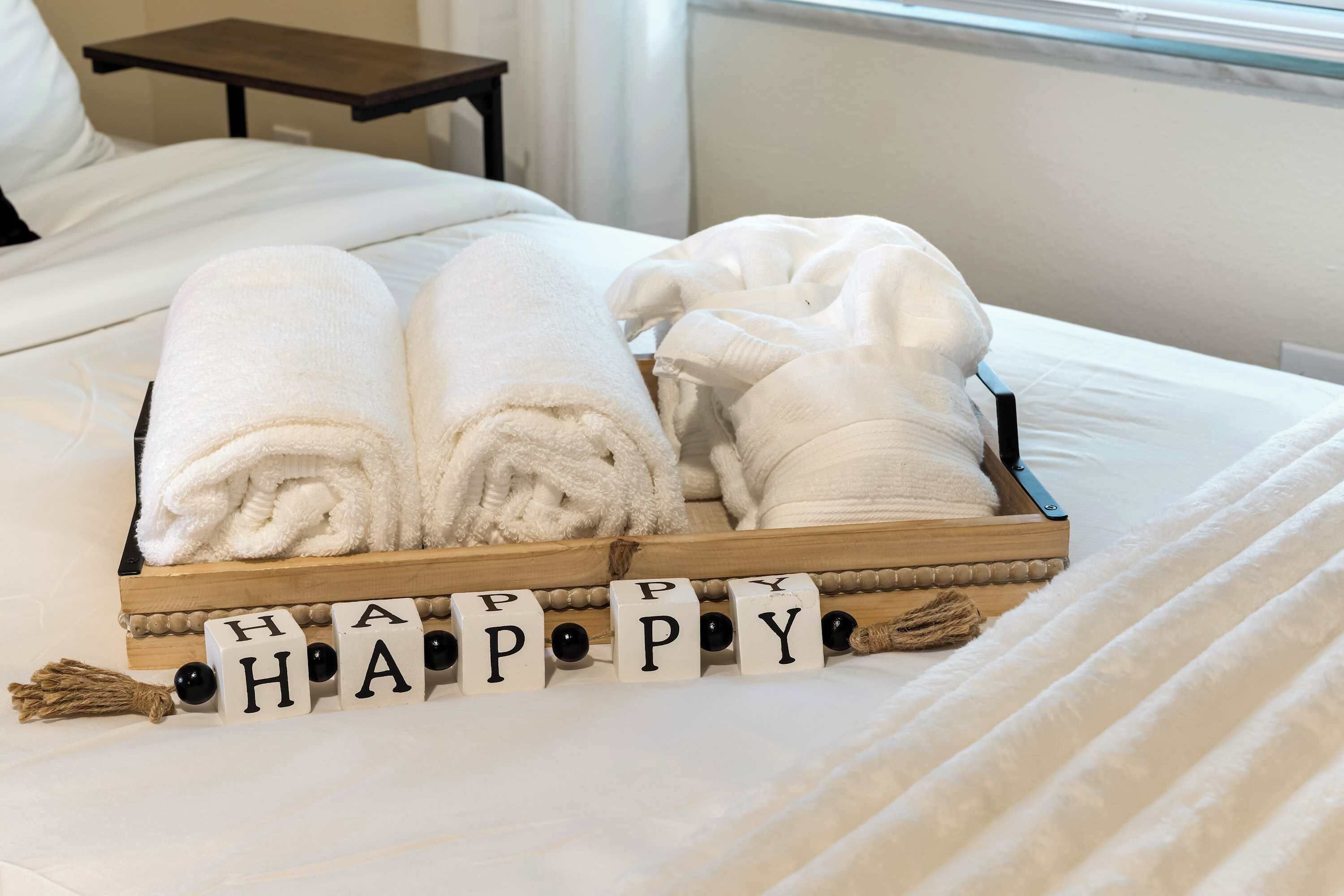 Una cama con toallas blancas ordenadas y una bandeja con la palabra 'HAPPY' escrita en bloques de madera.