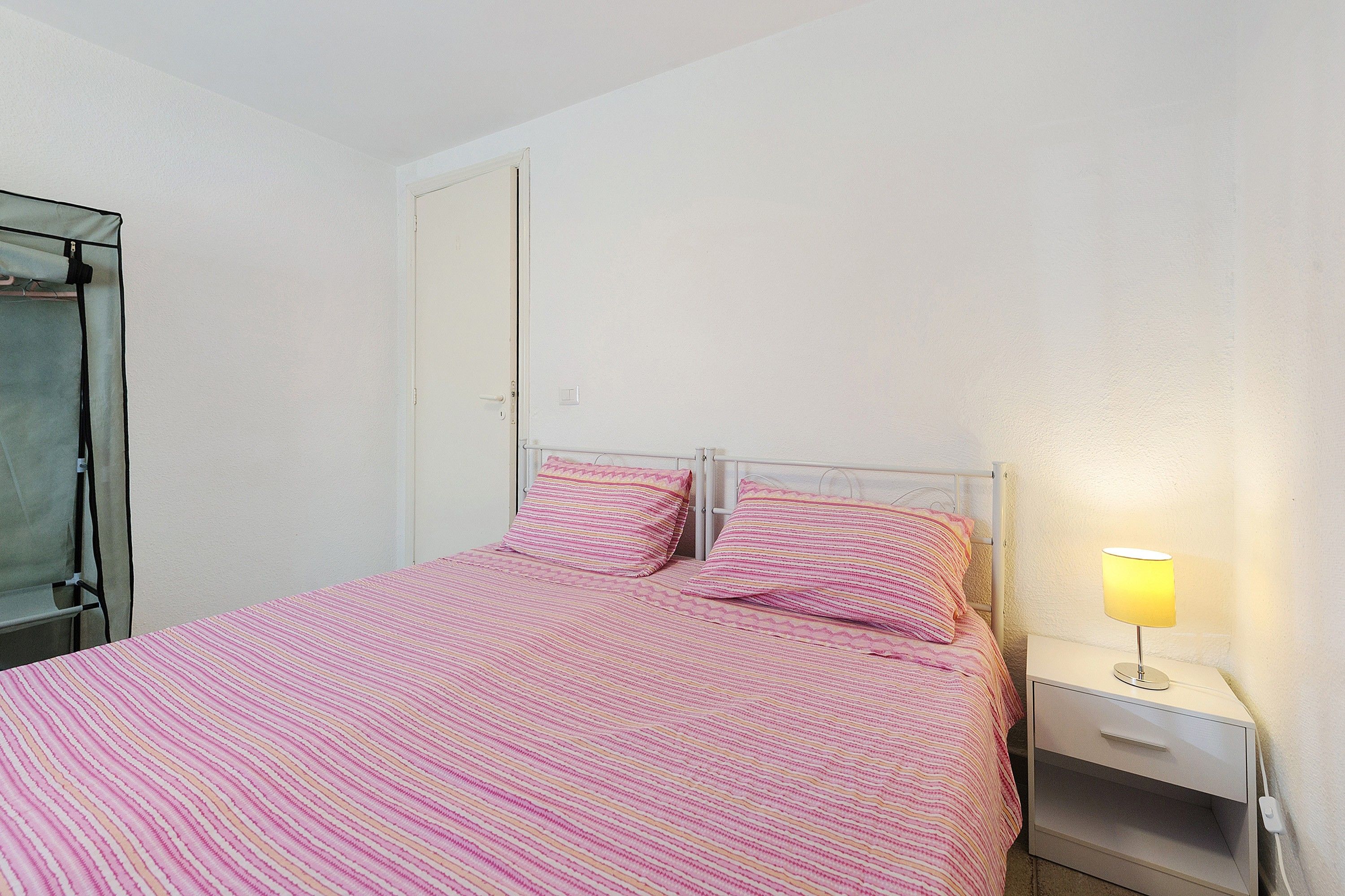 A single bed with a pink striped bedspread and matching pillows, a bedside table with a lamp, and a wardrobe with a mirror.