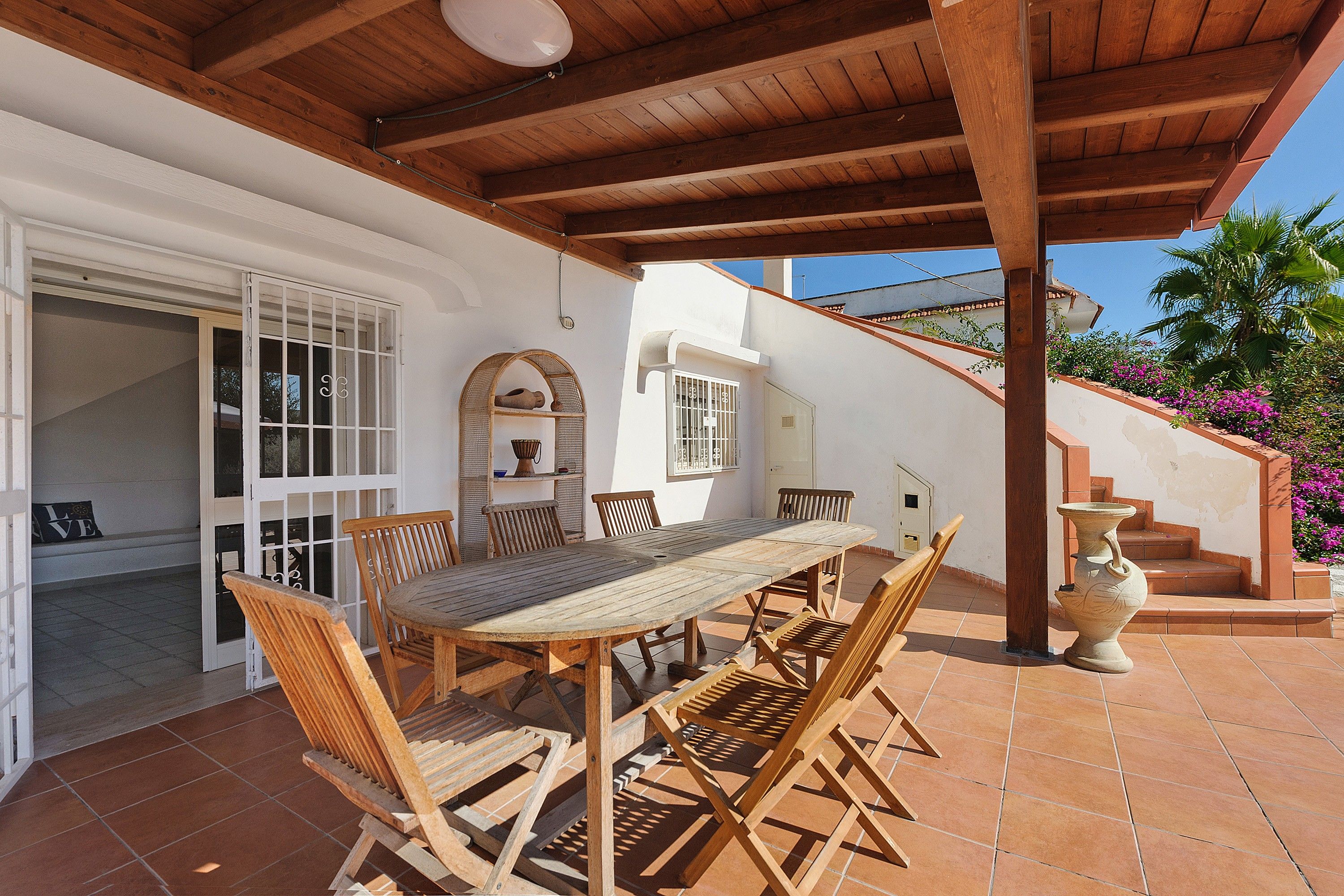 An outdoor patio with a wooden table and chairs, a white wall with a window, and a staircase leading to a higher level.