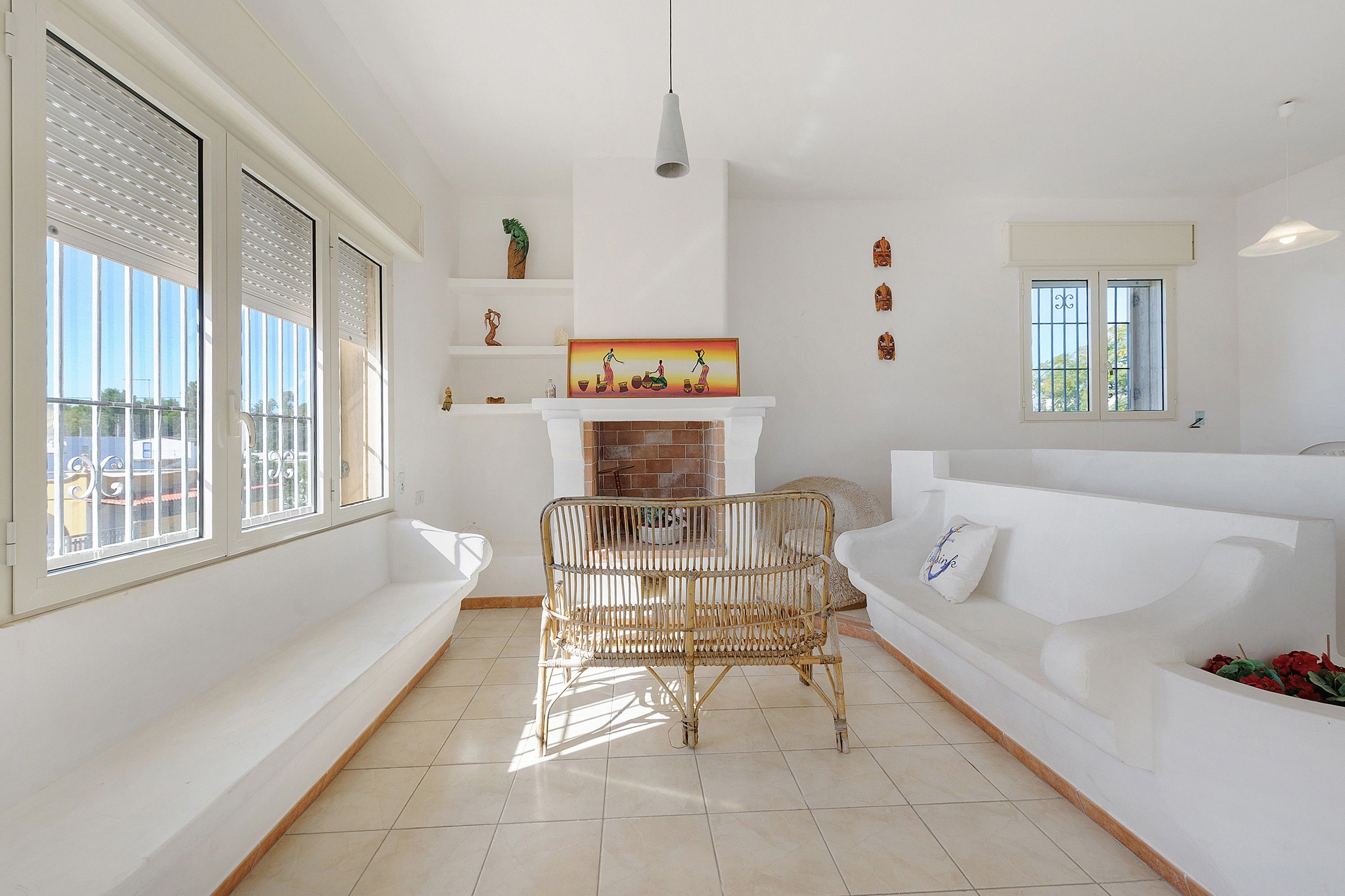 A spacious living room with a white sofa, a wicker chair, a fireplace, and a large window with blinds.