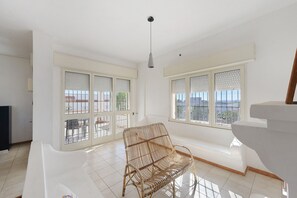 Quarto amplo com janelas grandes, uma cadeira de vime e teto branco.