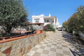 Rua residencial com uma casa branca, muros de pedra e um caminho pavimentado até a casa.