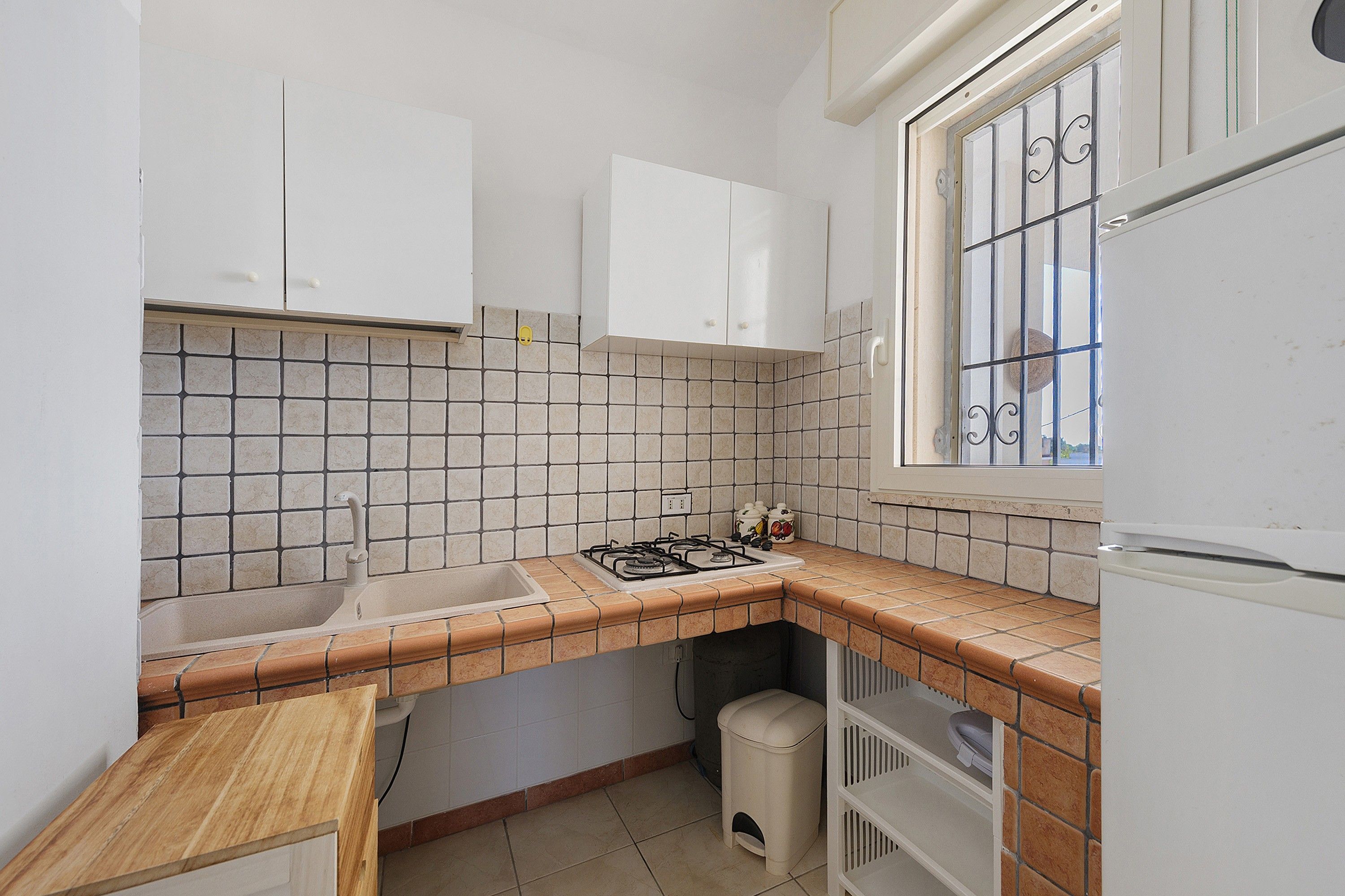 Een kleine keuken met witte keukenkastjes, een gasfornuis, een spoelbak en een raam met decoratieve roedeversiering.