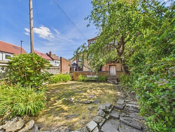 A residential backyard with a stone pathway, a wooden bench, and a variety of plants.