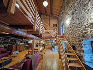 A rustic interior with wooden beams, a stone wall, a wooden staircase, and a seating area with a bench and a table.