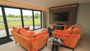 A modern living room with a large TV, orange sofas, and a view of a green landscape through large windows. - The Cozie Sow (Yeovil)