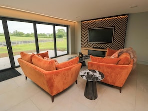 A modern living room with a large TV, orange sofas, and a view of a green landscape through large windows. - The Cozie Sow (Yeovil)