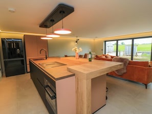 A modern kitchen with a central island, a dining area with a sofa, and a view of the outdoors. - The Cozie Sow (Yeovil)